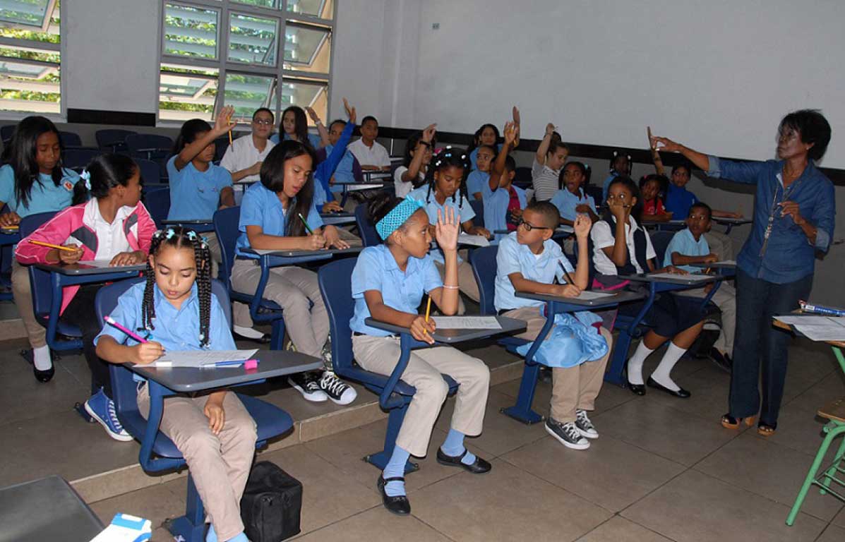  imagen Grupo de estudiantes en clase junto a su maestra, algunos levantan sus manos para participa. 